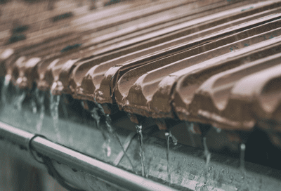 A close-up view of brown clay roof tiles during rainfall, with water streaming off the edge of the tiles and into a metal gutter below.