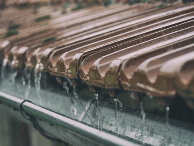 A close-up view of brown clay roof tiles during rainfall, with water streaming off the edge of the tiles and into a metal gutter below.