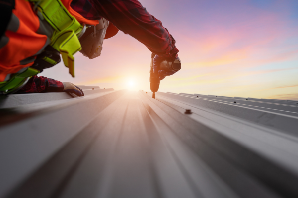 A close-up view of a worker in a red plaid shirt, reflective vest, gloves, and safety harness using a power drill to secure a screw into a metal roofing panel. The sun sets in the background under a pink and blue sky.