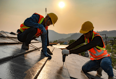 Two workers wearing yellow hard hats, safety glasses, and reflective orange vests kneel on a rooftop. One holds a power drill while the other steadies a panel. The sun sets over mountains and a village in the background.