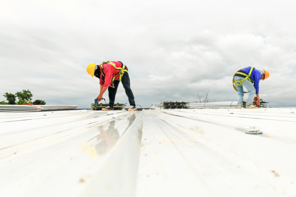 Two workers wearing hard hats and safety harnesses use power drills to install white metal roofing panels on a large flat rooftop. Stacks of additional panels sit beside them, and an overcast sky fills the background.