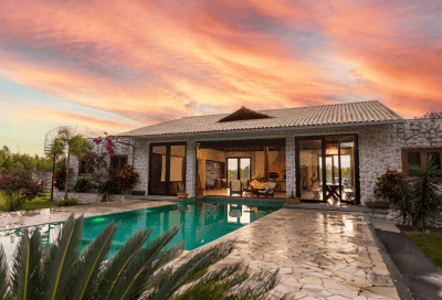 A single-story stone home with large glass doors and a peaked roof, featuring an outdoor pool and stone patio in the foreground. Palm plants and flowering bushes surround the property, with a pink and orange sunset sky in the background.