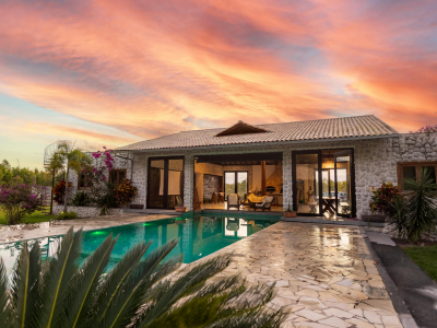 A single-story stone home with large glass doors and a peaked roof, featuring an outdoor pool and stone patio in the foreground. Palm plants and flowering bushes surround the property, with a pink and orange sunset sky in the background.