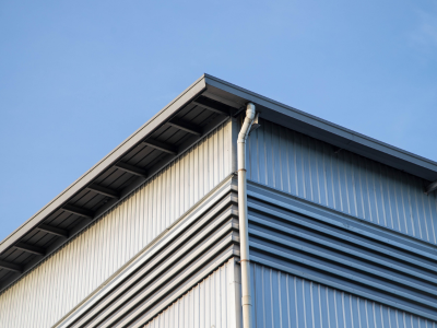 A close-up view of the upper corner of a commercial building with white and gray metal siding, dark metal roofing, and a white downspout running down the corner. A clear blue sky is visible in the background.