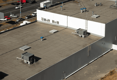 An aerial view of a large commercial building with a flat rooftop featuring HVAC units, roof vents, and a service ladder. The white and gray exterior walls are visible, along with a parking lot filled with vehicles in the background.
