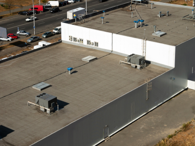 An aerial view of a large commercial building with a flat rooftop featuring HVAC units, roof vents, and a service ladder. The white and gray exterior walls are visible, along with a parking lot filled with vehicles in the background.