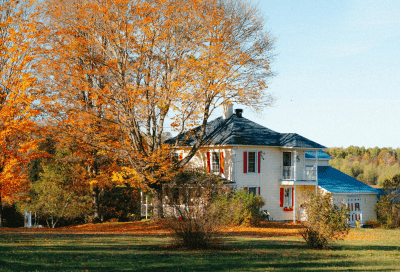 A cream-colored two-story home with red shutters and a covered porch, set among tall trees displaying vibrant orange and yellow fall foliage. A grass lawn scattered with fallen leaves is visible in the foreground.