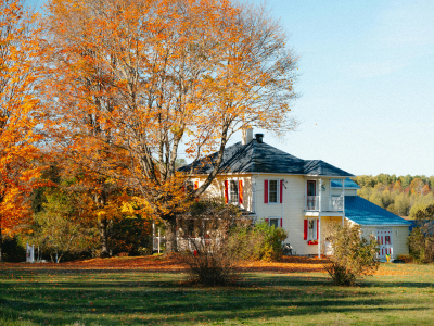 A cream-colored two-story home with red shutters and a covered porch, set among tall trees displaying vibrant orange and yellow fall foliage. A grass lawn scattered with fallen leaves is visible in the foreground.