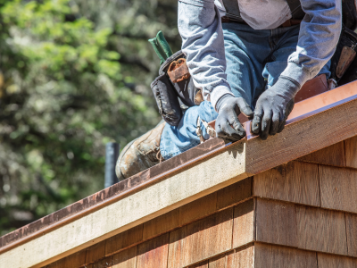 A close-up of a worker wearing dark work gloves and a tool belt, positioning copper flashing along the edge of a shingle roof. The blurred background shows greenery.
