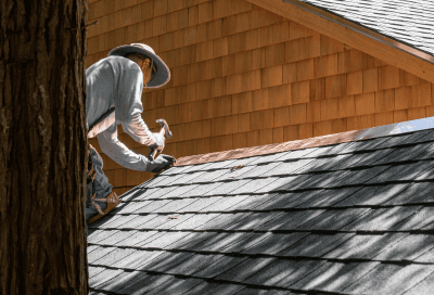 A person wearing a wide-brimmed hat and long sleeves kneels on a gray shingle roof, installing new shingles. The home features cedar shake siding, and a tree trunk is visible in the foreground.