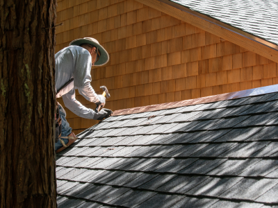 A person wearing a wide-brimmed hat and long sleeves kneels on a gray shingle roof, installing new shingles. The home features cedar shake siding, and a tree trunk is visible in the foreground.