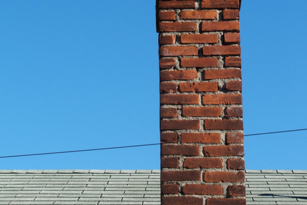 A red brick chimney rising through a gray asphalt shingle roof against a clear blue sky, showing the joint where the masonry meets the shingles.