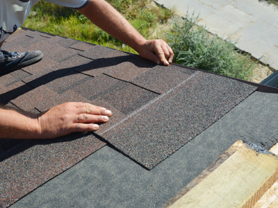 A roofer kneeling on a roof and positioning a new asphalt shingle by hand alongside the existing courses during a repair.
