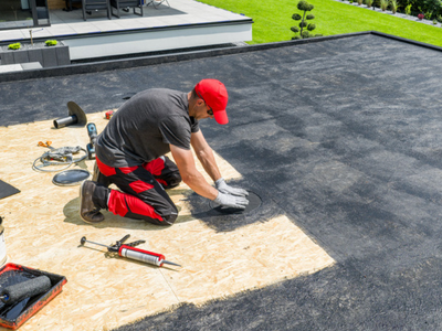 A roofer kneeling on a flat roof, pressing down a dark membrane patch beside board decking and sealing tools.