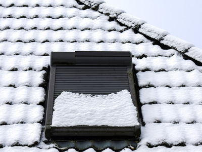 A tile roof covered in fresh snow with a dark skylight and closed roller shutter, showing winter conditions around a roof penetration.